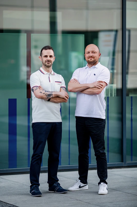 Stojící portrét dvou mužů v bílých polo tričkách stojících před moderní prosklenou budovou.