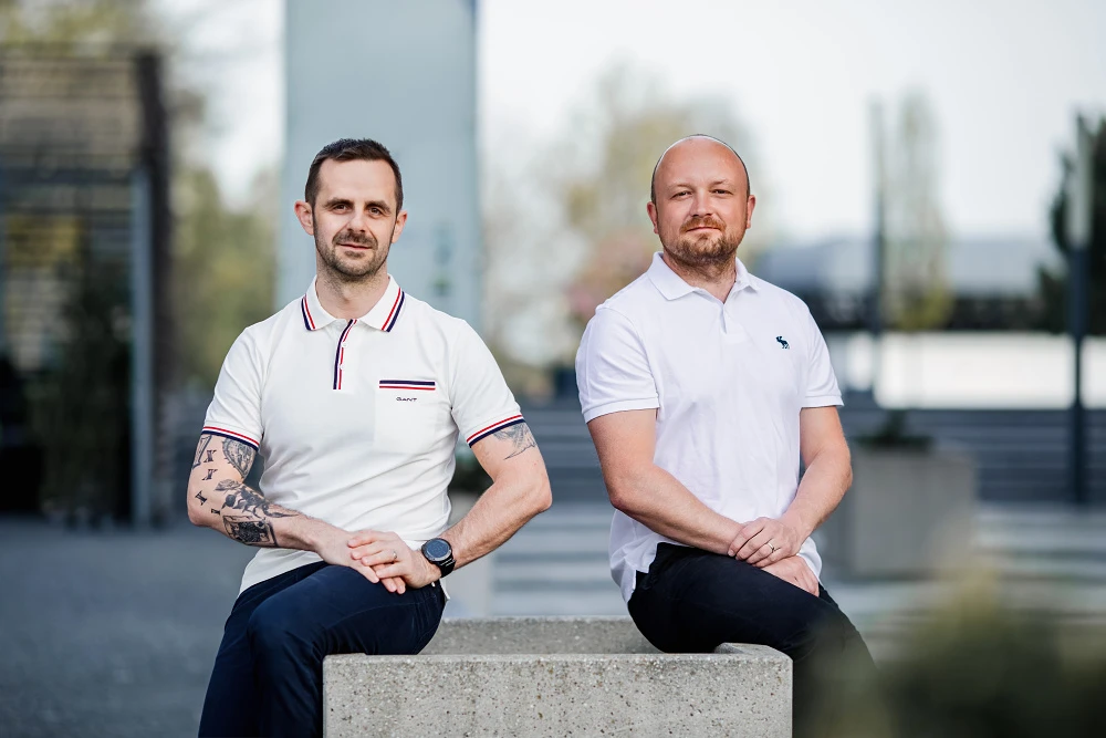 Uvolněný portrét dvou kolegů sedících v exteriéru, styl business casual, rozostřené městské pozadí.