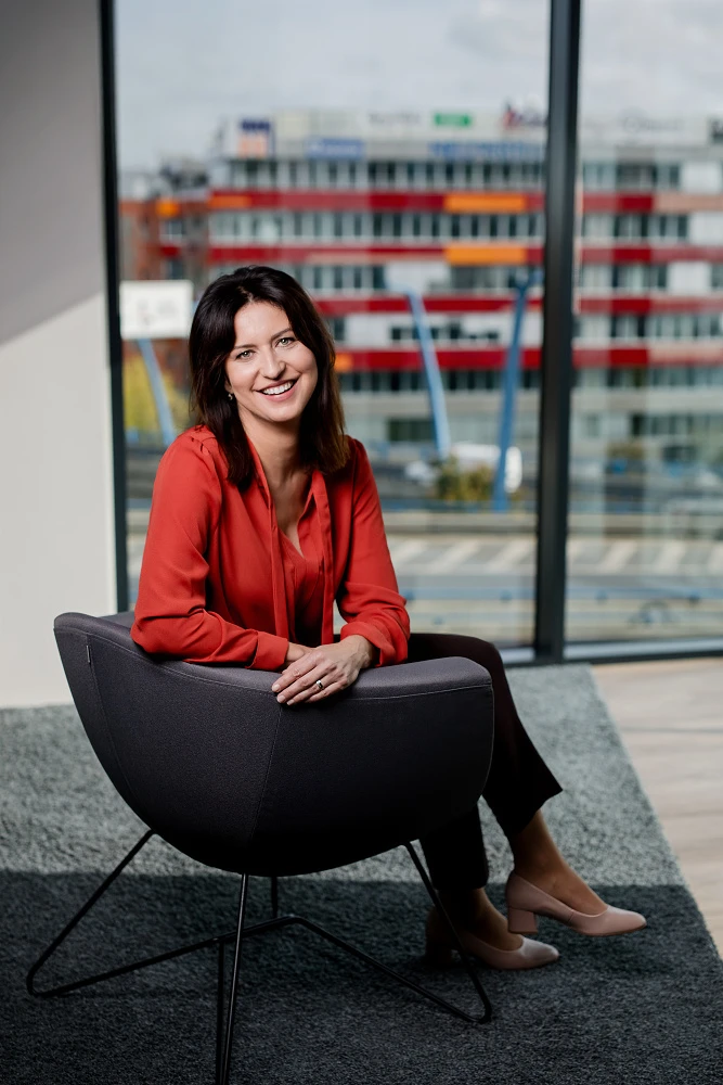 Portrét usmívající se manažerky v červené halence sedící v moderním křesle s výhledem z kanceláře.