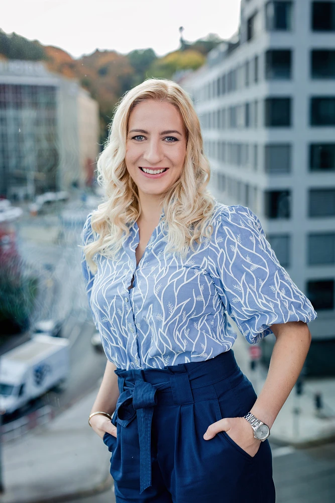 Portrét ženy v modré vzorované halence v moderním kancelářském prostředí s rozostřeným pozadím města.