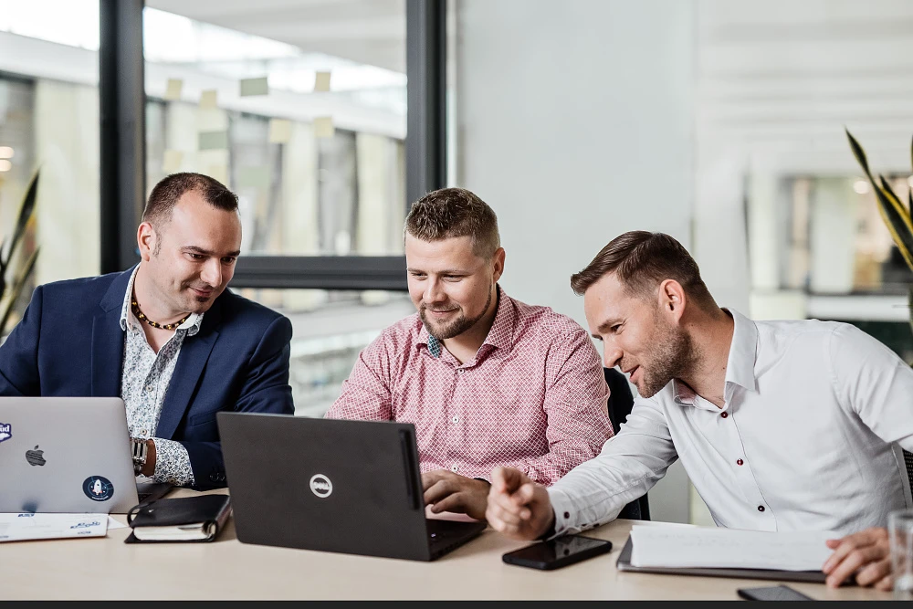 Reportážní fotografie týmu tří kolegů diskutujících nad prací u notebooku v prosvětlené kanceláři.