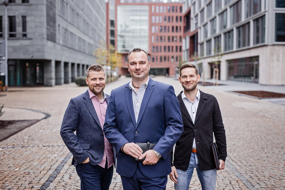 Reprezentativní skupinový portrét tří mužů v byznys oblečení před moderní prosklenou budovou.